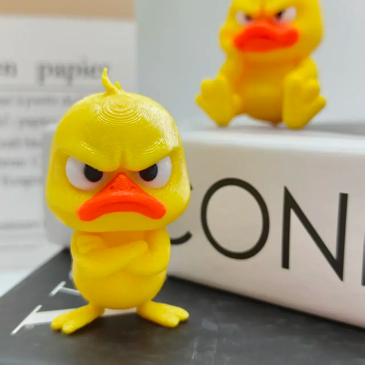A yellow 3D printed angry duck figurine with orange beak sitting on a dark surface.