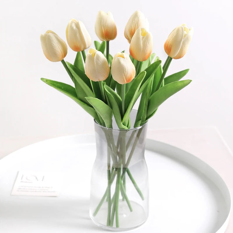 A bouquet of light yellow artificial tulips in a clear glass vase on a white plate.