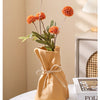 A terracotta money bag shaped vase holds dried orange flowers and green leaves.