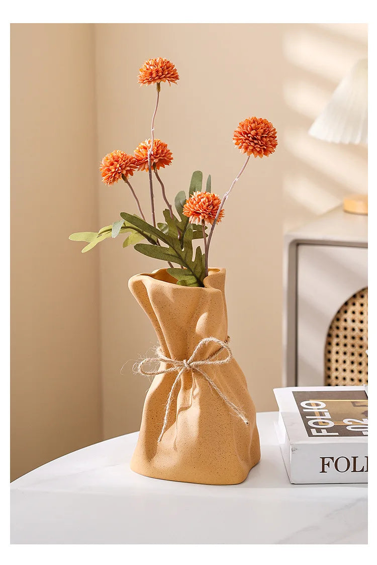A terracotta money bag shaped vase holds dried orange flowers and green leaves.