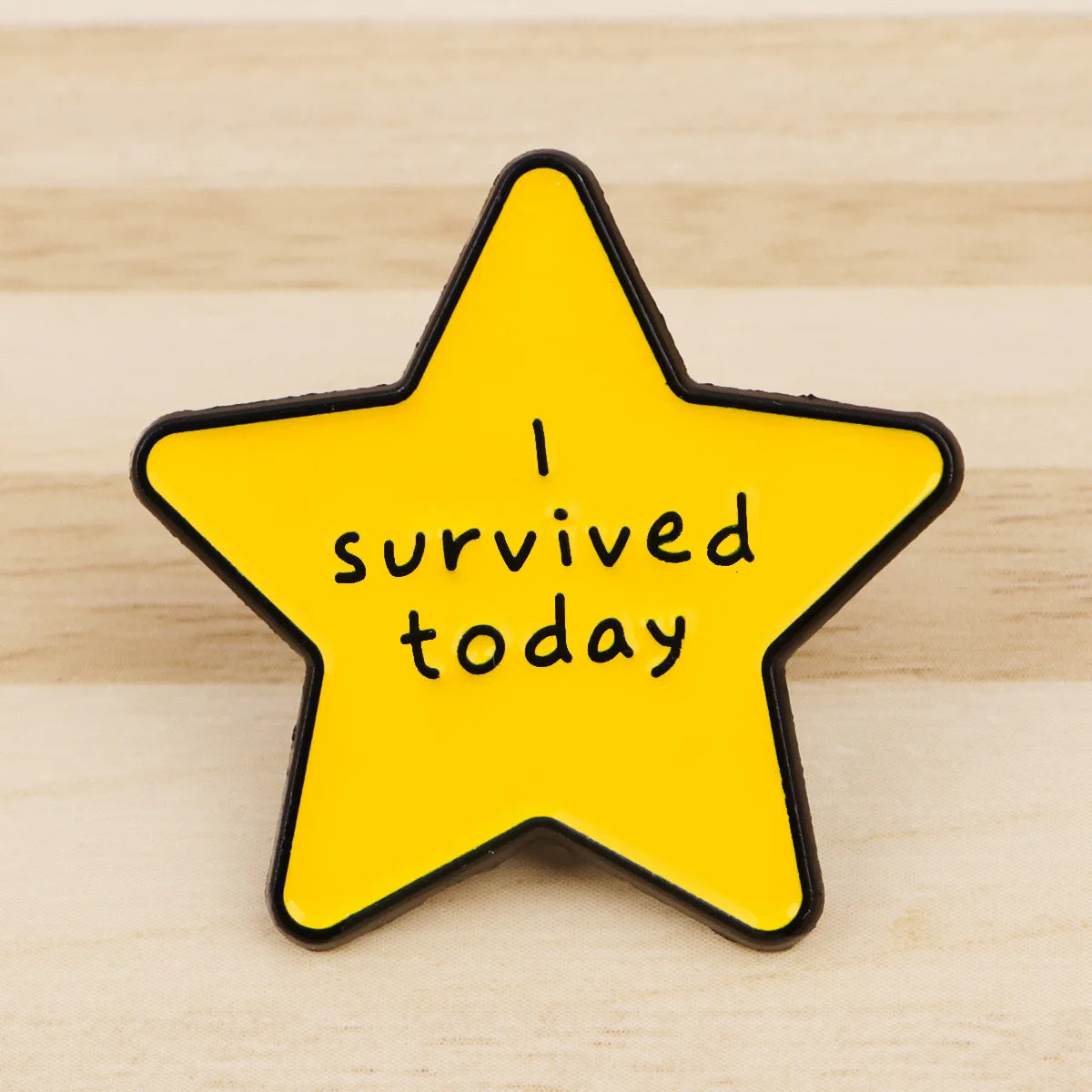 A yellow star enamel pin with black outline featuring the text I survived today.