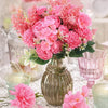 A bouquet of pink and coral artificial silk peony flowers in a gold vase on a white lace tablecloth.