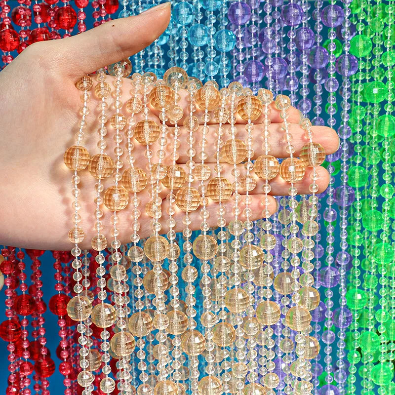 Une main tient plusieurs brins de perles d'ambre et de cristal clair sur un fond de rideau de perles coloré.
