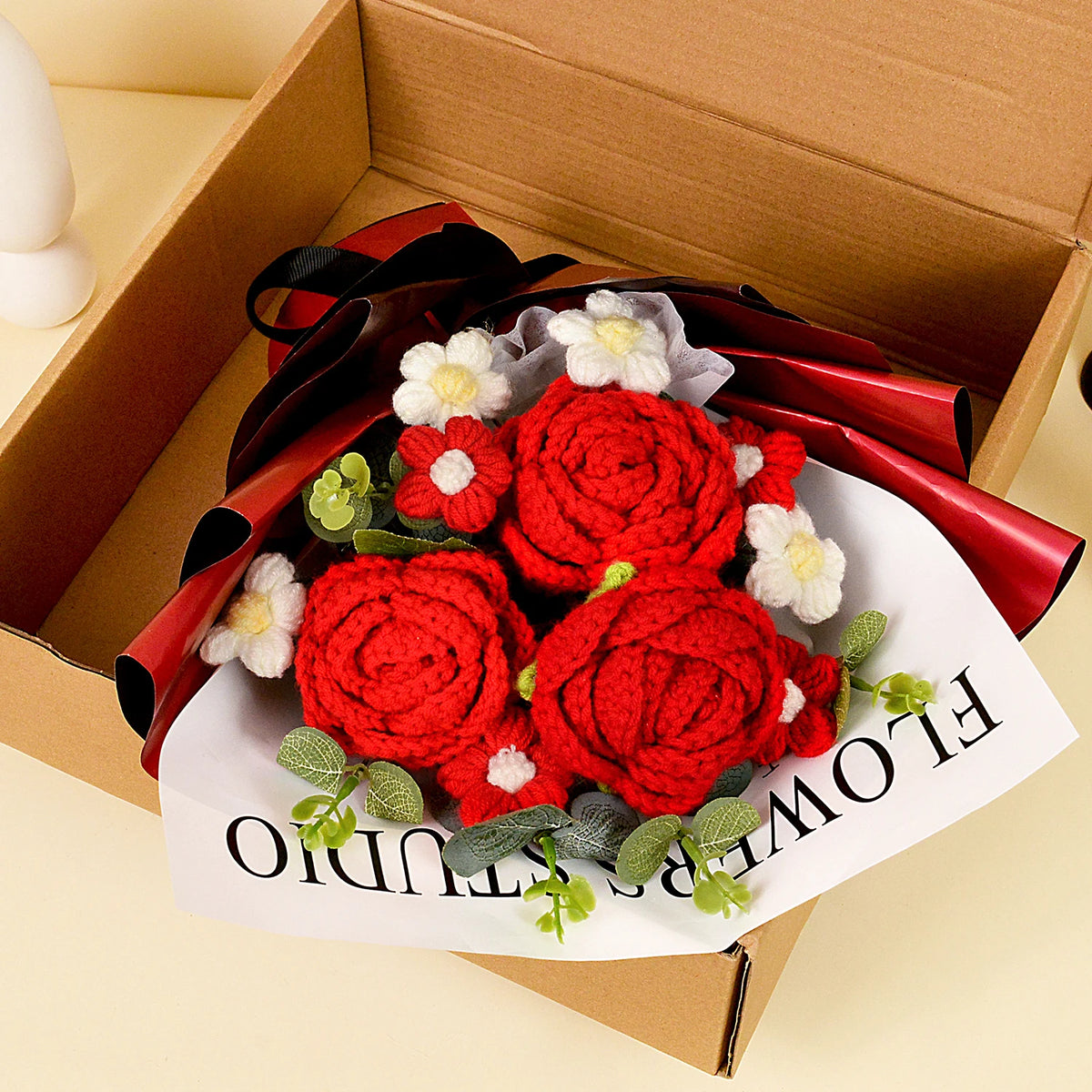 A bouquet of handcrafted red crochet roses with white accents is presented in a red ribbon inside a cardboard box.
