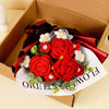 A bouquet of handcrafted red crochet roses with white accents is presented in a red ribbon inside a cardboard box.
