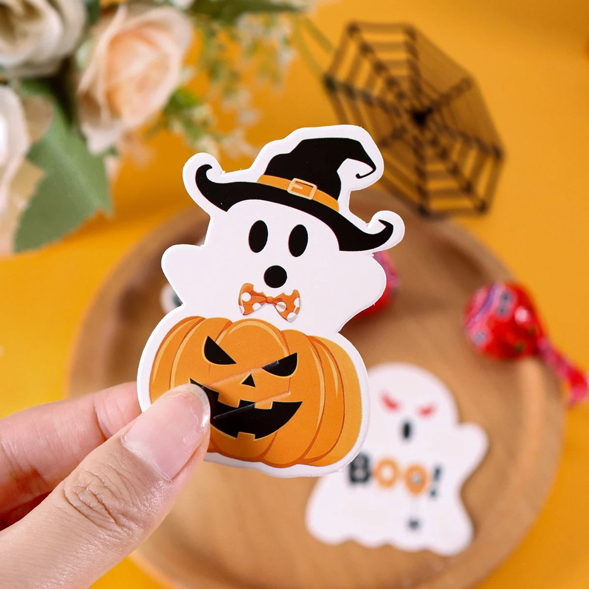 A hand holds an orange pumpkin ghost pattern lollipop holder card against a yellow background.