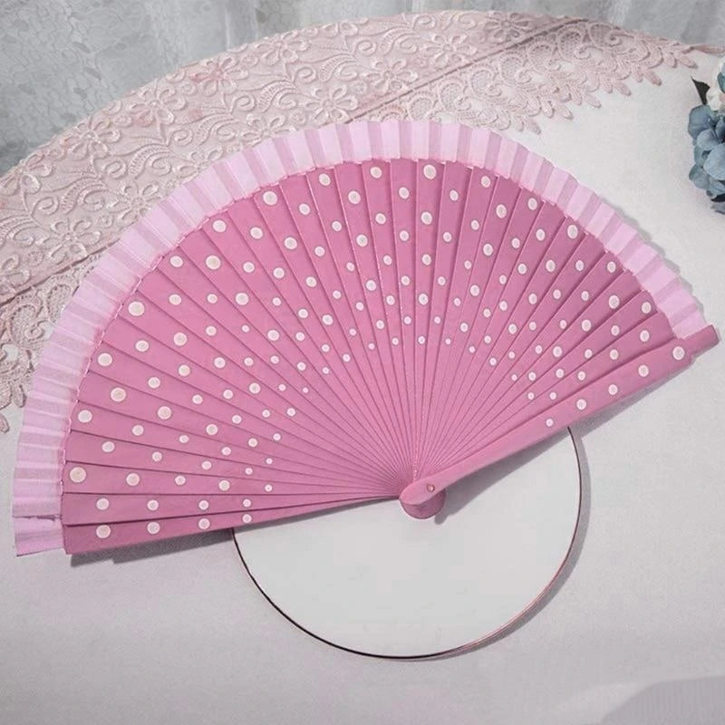 A pink hand fan with white polka dots is displayed on a white cloth next to a lace fabric.