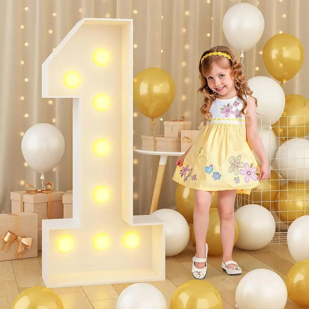A large light-up number one decoration made of LED lights stands next to a young girl surrounded by yellow and white balloons