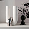 White ceramic U-shaped tabletop vase and black spherical sculpture displayed on a white shelf.