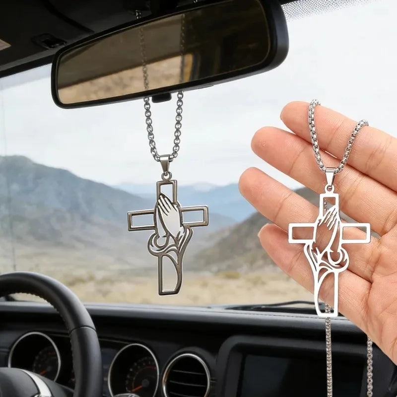 A hand holds a stainless steel praying hands cross pendant hanging from a chain, positioned in front of a car mirror.