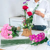 A person holds a bouquet of red and pink roses and greenery on a white table, with green floral wire stems laid out nearby.