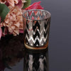 Elegant glass votive candle holder with a black and silver chevron pattern, set against a blurred floral background.