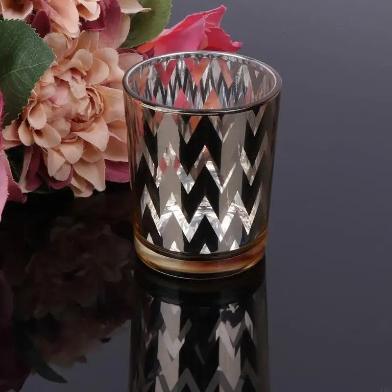 Elegant glass votive candle holder with a black and silver chevron pattern, set against a blurred floral background.