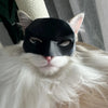 A white cat wearing a black bat mask with its tongue sticking out.