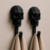A pair of black 3D skull wall hooks mounted on a wall, with two beige towels hanging from them.