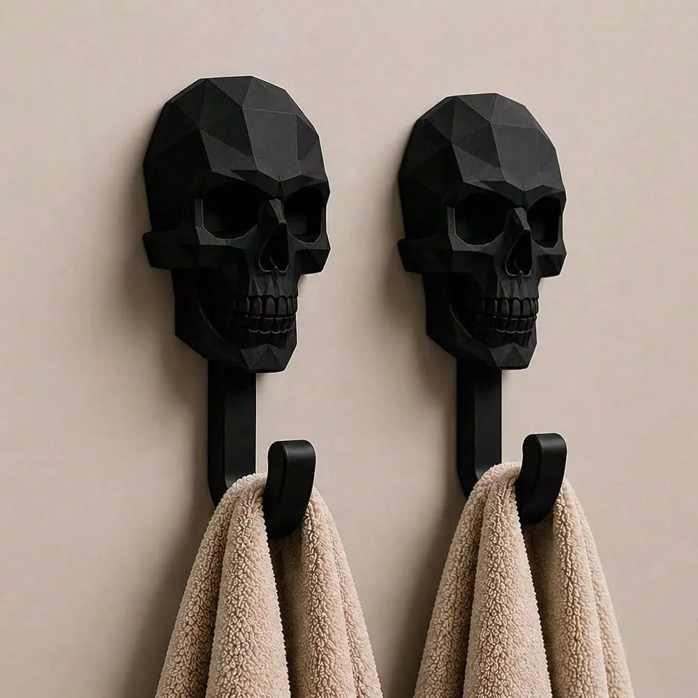 A pair of black 3D skull wall hooks mounted on a wall, with two beige towels hanging from them.
