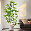 An artificial bamboo branch with green leaves stands in a white pot next to a brown sofa.