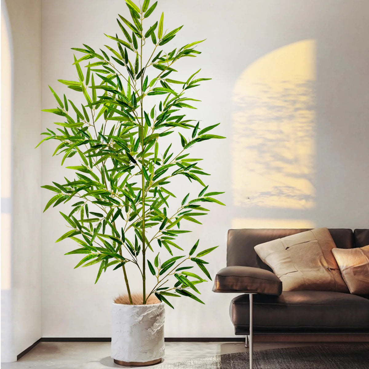 An artificial bamboo branch with green leaves stands in a white pot next to a brown sofa.