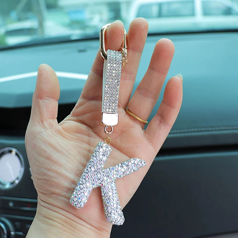 A hand holds a crystal letter keychain charm with a gold loop and a sparkling letter on a dark car surface.