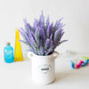 A white ceramic vase filled with purple lavender flowers sits on a white surface next to a yellow bottle and a blue glass.