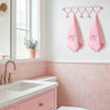 A pink heart over-the-door hook rack with five pink fabric hooks displayed on a white wall above a pink tiled vanity.