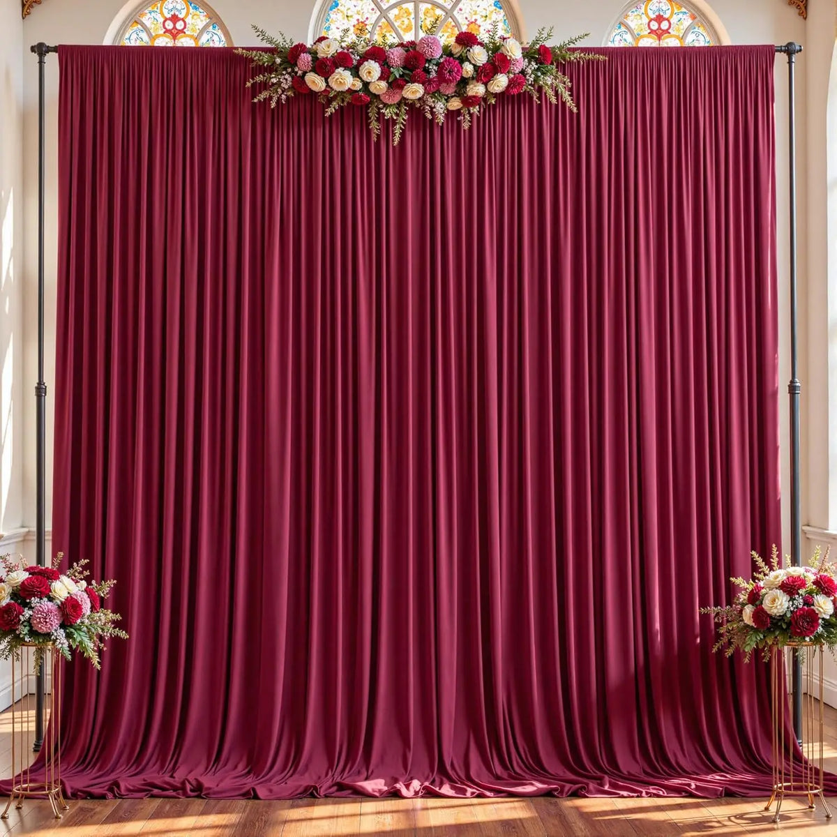 A deep burgundy fabric backdrop with gathered pleats and decorated with floral arrangements for an event.