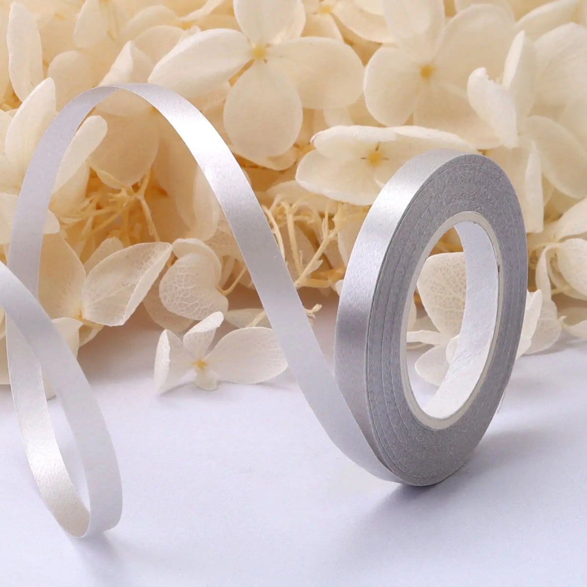 A roll of silver satin balloon ribbon unwinding against a background of light cream flowers.