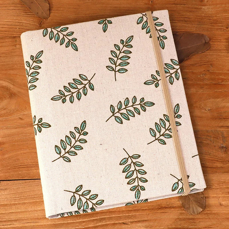 A cream linen photo album with a repeating pattern of green leaves and a beige ribbon on a wooden surface.