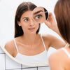 A woman uses a magnifying compact mirror with a suction cup to examine her eye.