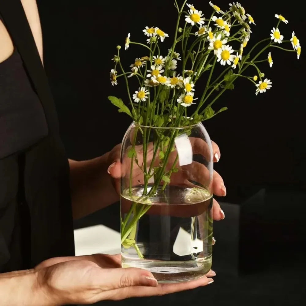 A hand holds a clear glass hydroponic vase filled with green plants and yellow daisy flowers against a dark background.