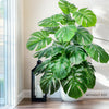 A large artificial monstera plant with green silk leaves displayed indoors near a window.