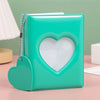 A teal heart-shaped window photocard album with a silver chain detail resting on a pink surface.