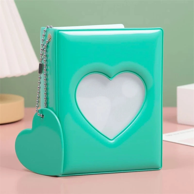 A teal heart-shaped window photocard album with a silver chain detail resting on a pink surface.