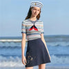 A woman wearing a striped shirt, dark skirt, and a captain's hat with a red scarf by the sea.