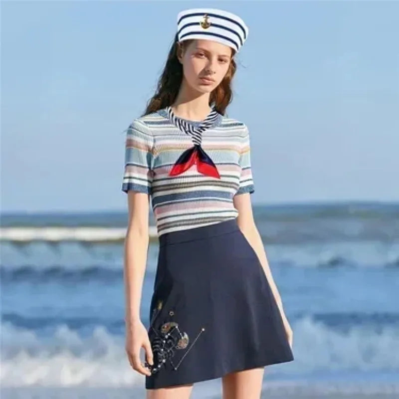 A woman wearing a striped shirt, dark skirt, and a captain's hat with a red scarf by the sea.
