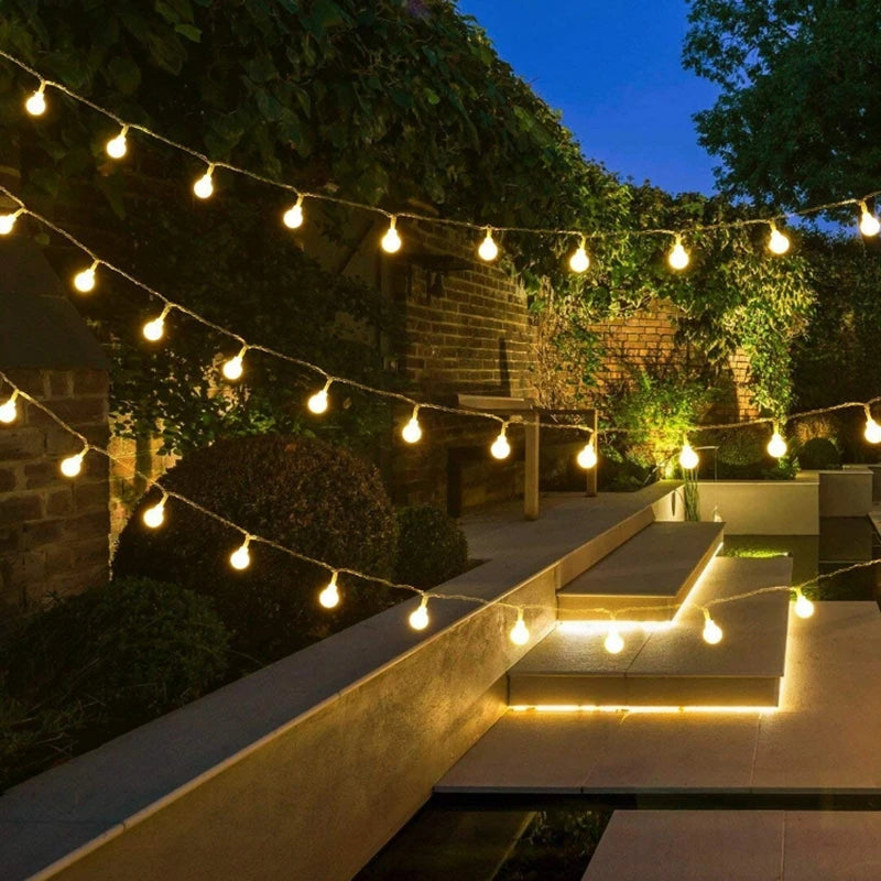 String lights illuminate a garden pathway and stone wall at dusk.