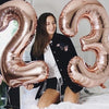 A young woman is sitting on a bed surrounded by large rose gold foil balloons shaped like the number 23.
