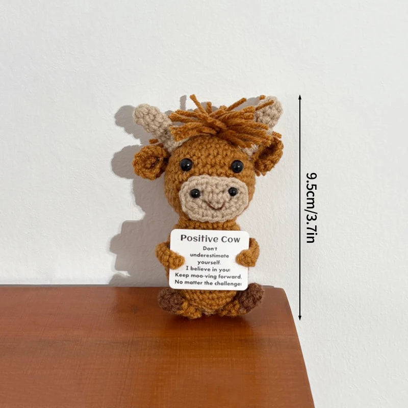 A handmade crochet positive cow doll with a slogan on its tag displayed against a white wall.