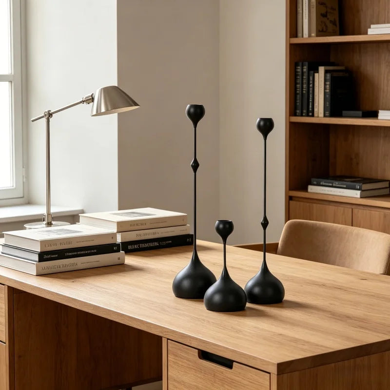 A set of three matte black waterdrop design candle holders displayed on a wooden table next to a desk lamp.