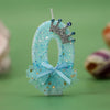 A light blue glitter candle shaped like the number 0 decorated with a silver crown and a sheer blue ribbon.