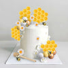 A white cake decorated with yellow honeycomb shapes, small white and gold flowers, and a bee-themed topper set.