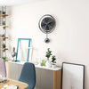 A modern circular wall clock with black face and Arabic numerals hangs on a white wall above a desk area.