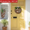 A round wooden welcome sign with greenery and a bow hanging on a yellow door.