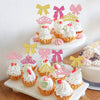 A display of elegant crown and bow cupcake toppers in pink, gold, and white on a white cake stand.