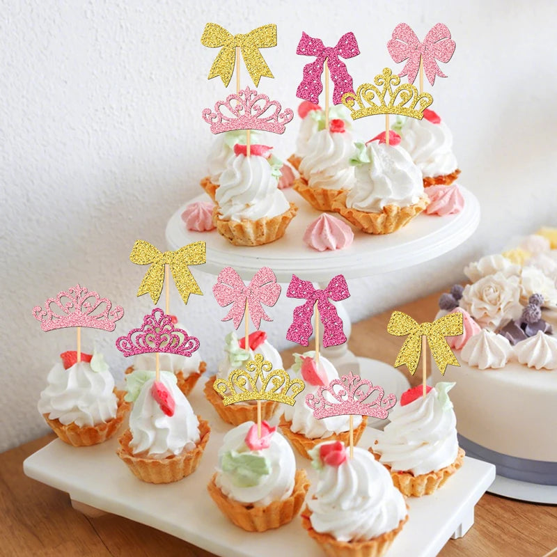 A display of elegant crown and bow cupcake toppers in pink, gold, and white on a white cake stand.