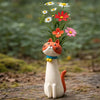 A whimsical resin cat figurine with orange and white fur, holding a bouquet of colorful flowers.