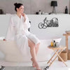 A woman sits in a white bathtub in a modern bathroom with an elegant iron sign on the wall.