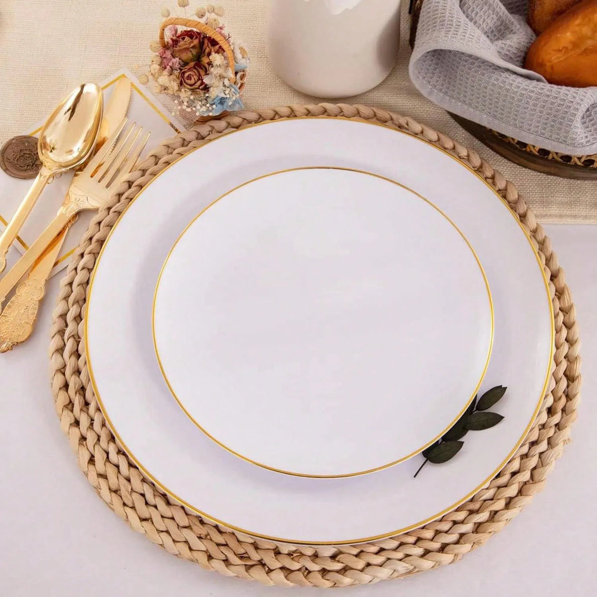 A white plate with a gold rim is set on a woven placemat next to gold cutlery and a small floral arrangement.