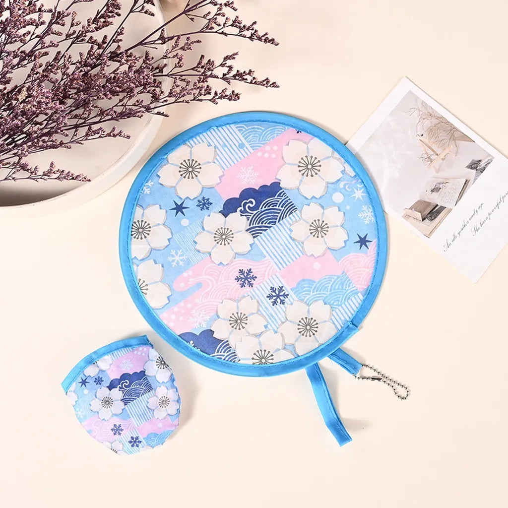 A round, light blue fabric fan with pink and white floral and geometric patterns is shown next to a dried flower branch.