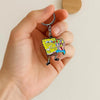 A hand holds a cartoon keychain featuring SpongeBob SquarePants in yellow and pink colors.